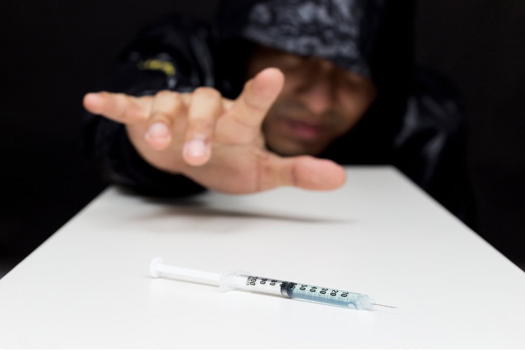 A person in a hooded sweatshirt reaches out towards a syringe on a white table, symbolizing the struggle and temptation associated with addiction. This moment reflects the challenging path to recovery found in Fentanyl rehab in Reseda CA.
